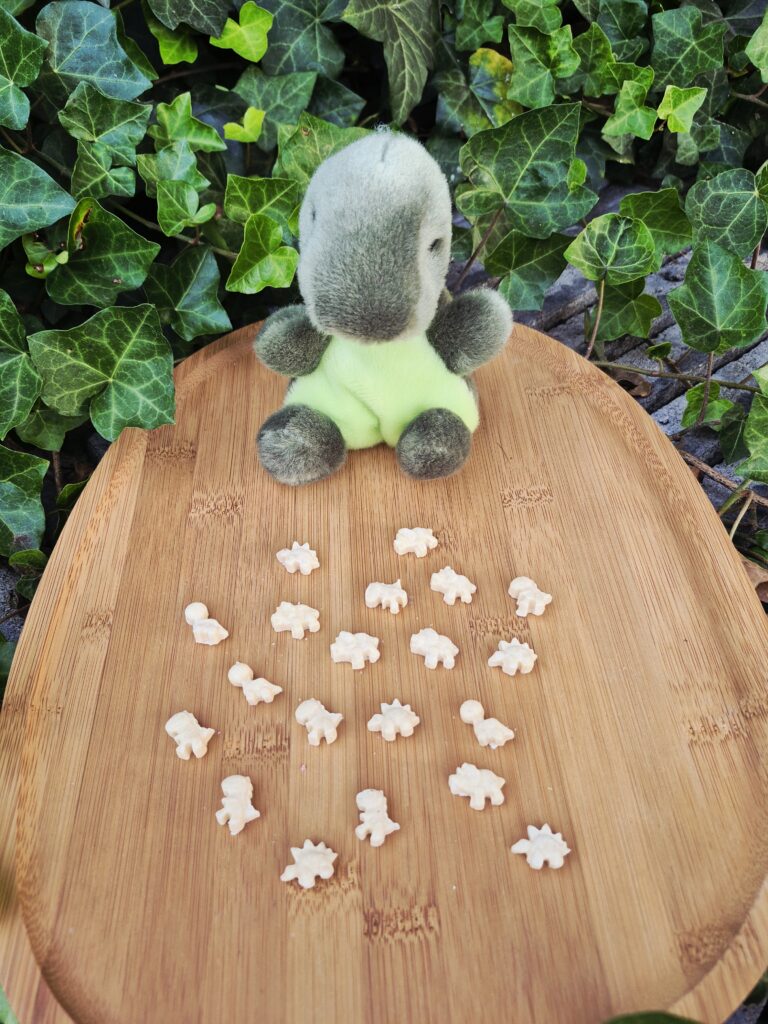 Mini DinoPawrs hondenkoekjes in verschillende dinosaurusvormen, gepresenteerd op een houten plank naast een schattige dinosaurusknuffel. De kleine koekjes zijn ideaal als trainingssnoepje, beloning of tussendoortje voor honden.