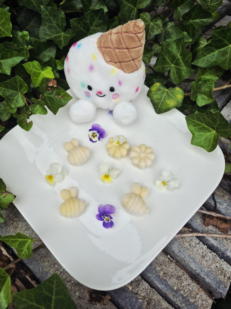 Gelato Scoops Naturel hondenijsjes op basis van geitenmelk in de vorm van bijtjes en bloemetjes, gepresenteerd op een wit bord met eetbare viooltjes en een vrolijke ijsjesknuffel op de achtergrond.