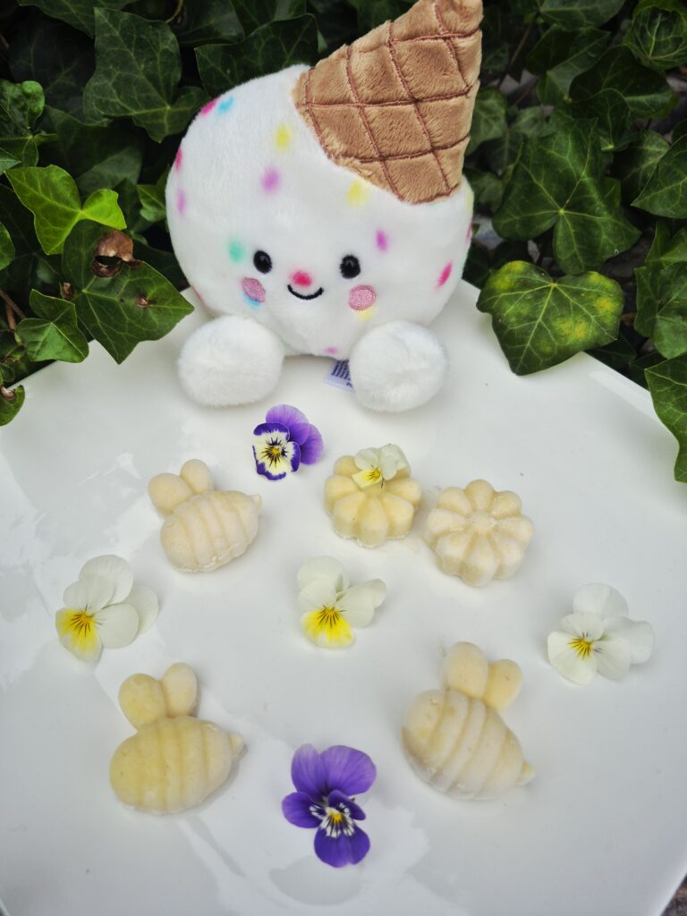 Gelato Scoops Appel hondenijsjes op basis van geitenmelk en appel, gegoten in schattige bij- en bloemvormen en gepresenteerd op een wit bord met eetbare viooltjes en een vrolijke ijsjesknuffel op de achtergrond.