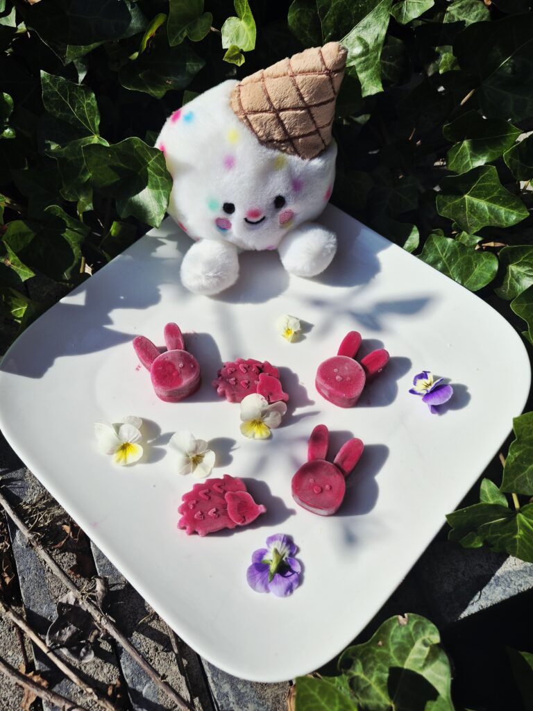 Gelato Scoops Purple Carrot hondenijsjes op basis van geitenmelk en paarse wortel, gegoten in schattige konijnen- en egelvormen en gepresenteerd op een wit bord met eetbare viooltjes en een vrolijke ijsjesknuffel op de achtergrond.